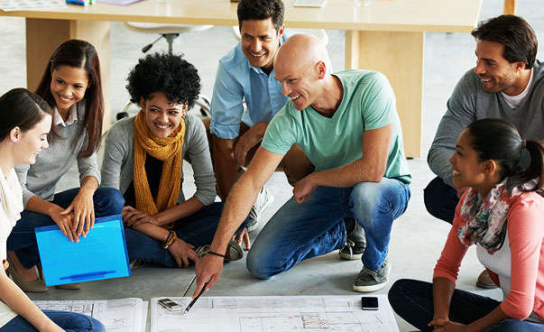 The picture shows a workshop-situation where a man shows the participants a poster which is lying on the floor.