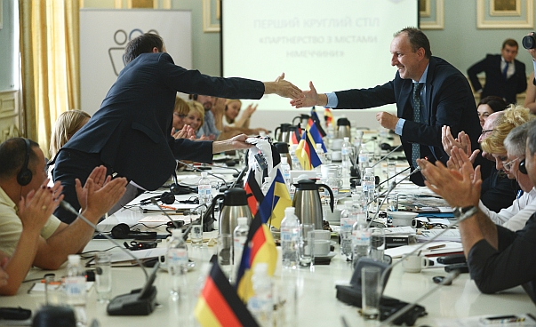 One German man and two Brazilian men and a Brazilian woman are sitting at e desk for a podium-discussion. The flags of Brazil and Germany are on the desk.